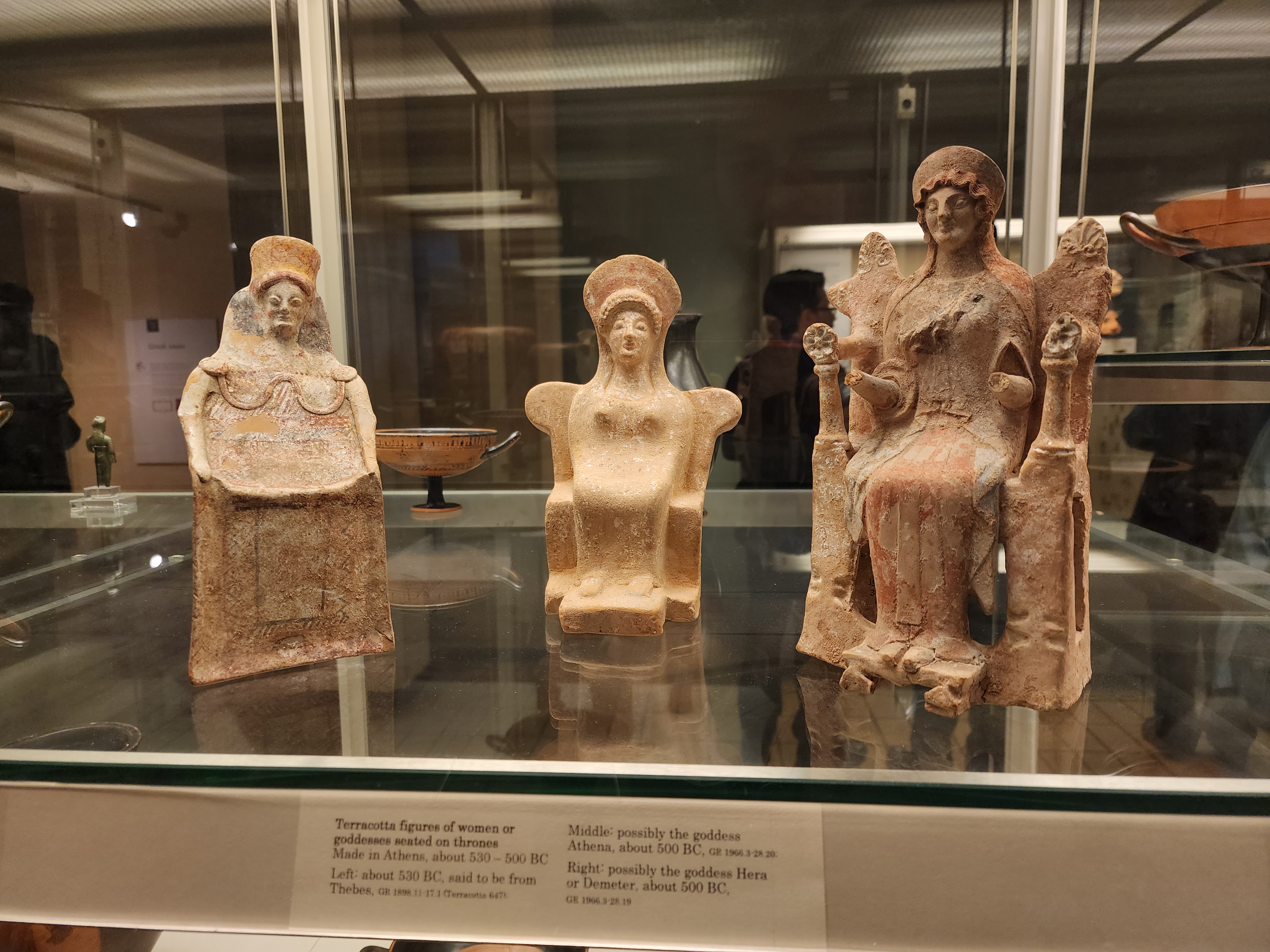 Three terracotta figurines of goddesses sitting on thrones, on a shelf in the British Museum